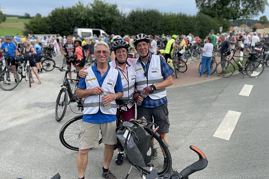 NRW-Radtag 2023 Die drei ADFC-Scouts mit Flaschen in der Hand stehen vor einer Menge an Radfahrerinnen und Radfahrern.
