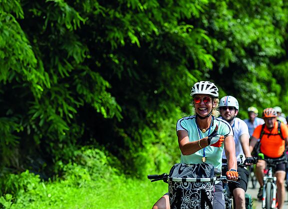 Eine Gruppe von Radfahrenden auf einer Straße entlang eines Waldes.