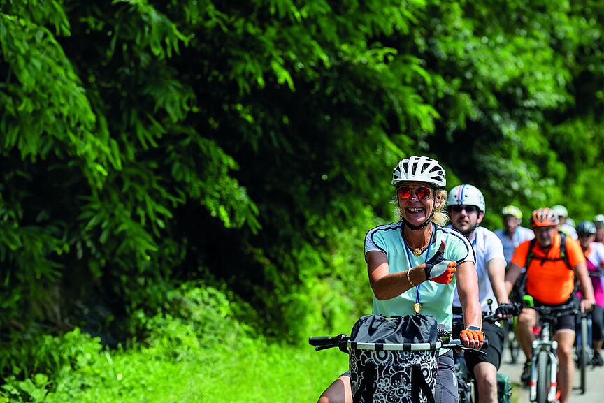 NRW Radtour 2025 Eine Gruppe von Radfahrenden auf einer Straße entlang eines Waldes.